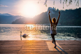 Naklejki Sun Salute Yoga. Young woman doing yoga by the lake at sunset, swan passing by