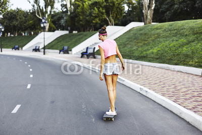Beautiful hipster girl in a jeans shorts and pink shirt walking with skateboard near the green park, sexy style, adult, long legs, brunette, white shoes, sexy model, posing on camera