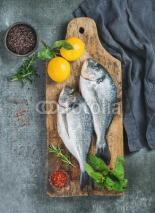 Fresh uncooked sea bream or dorado fish with lemon, herbs and spices in bowls on rustic wooden board over grey concrete background, top view. Healthy, dieting, clean eating concept