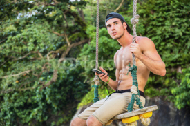 Shirtless athlete sitting with the smartphone on the swing. Horizontal outdoors shot. 