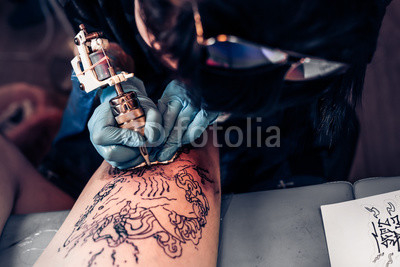 Closeup tattoo process in a professional salon. Master makes tattoo pattern on the client leg. Master works in sterile blue gloves.