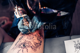 Naklejki Closeup tattoo process in a professional salon. Master makes tattoo pattern on the client leg. Master works in sterile blue gloves.