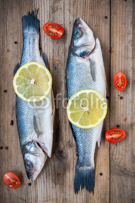 Two raw seabass fish with lemon and cherry tomatoes on wooden ba