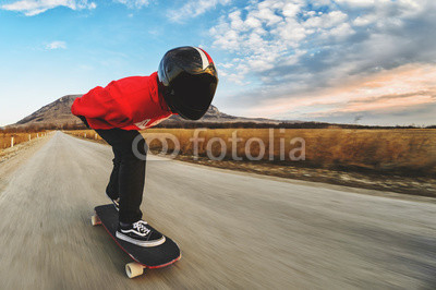 A young man in helmet and a leather suit in a special rack rides a longboard on afsaltu on background mountains and beautiful sky