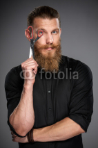 Serious hipster man with beard and mustashes holding scissors looking through it, over grey background