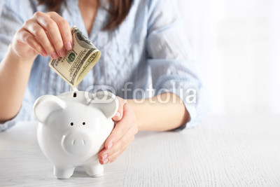 Woman sitting at table and putting money into piggy bank closeup