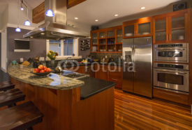 Contemporary upscale home kitchen interior with wood cabinets and floors, granite countertop, accent lighting, television & stainless steel appliances including double oven, refrigerator and vent hood