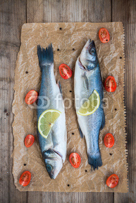 Two raw seabass fish with a lemon slice and cherry tomatoes on w