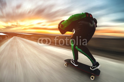 A man in a helmet and leather suit, in a rack at high speed, rides on a long longboard for downhill on afsalt against the backdrop of the setting sun in the light of the setting sun.